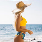 Woman in yellow bikini top and blue shorts with a straw hat on a beach.