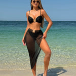 A model wearing a three-piece swimsuit set, which includes a bikini top, bikini bottoms, and a long mesh cover-up dress. The swimsuit is available in various colors and is shown in a beach setting.