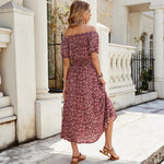 A full-length view of a person wearing a wine red, floral-printed, one-shoulder midi dress. The dress has a pleated A-line silhouette, an elastic waist, and a single short sleeve.