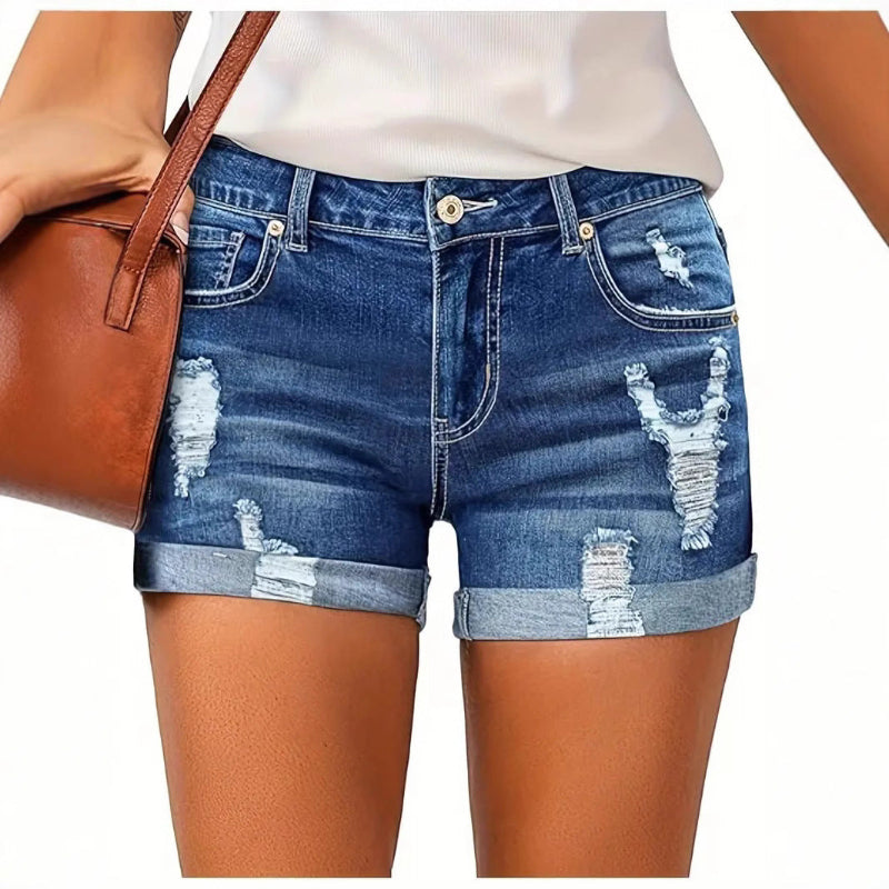 A model wearing deep blue, high-waisted denim shorts with a cuffed hem, paired with a white t-shirt. The frayed edges of the shorts are visible, and the overall style is casual and chic.