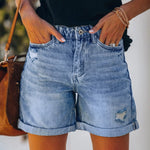 A pair of blue, high-waisted fringed denim shorts with distressed details. The shorts are laid flat on a neutral background, showcasing the style and texture. The fringe detailing is prominent along the hemline.