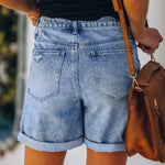 A pair of blue, high-waisted fringed denim shorts with distressed details. The shorts are laid flat on a neutral background, showcasing the style and texture. The fringe detailing is prominent along the hemline.