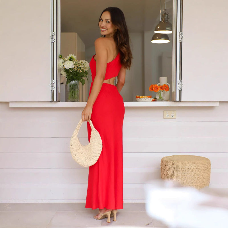 A woman wearing a sleeveless, solid red midi dress with an oblique collar. The dress has a long, flowing skirt, and she is standing in a resort-style setting.
