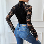 Person wearing a black lace long-sleeve top and blue jeans against a white wall.