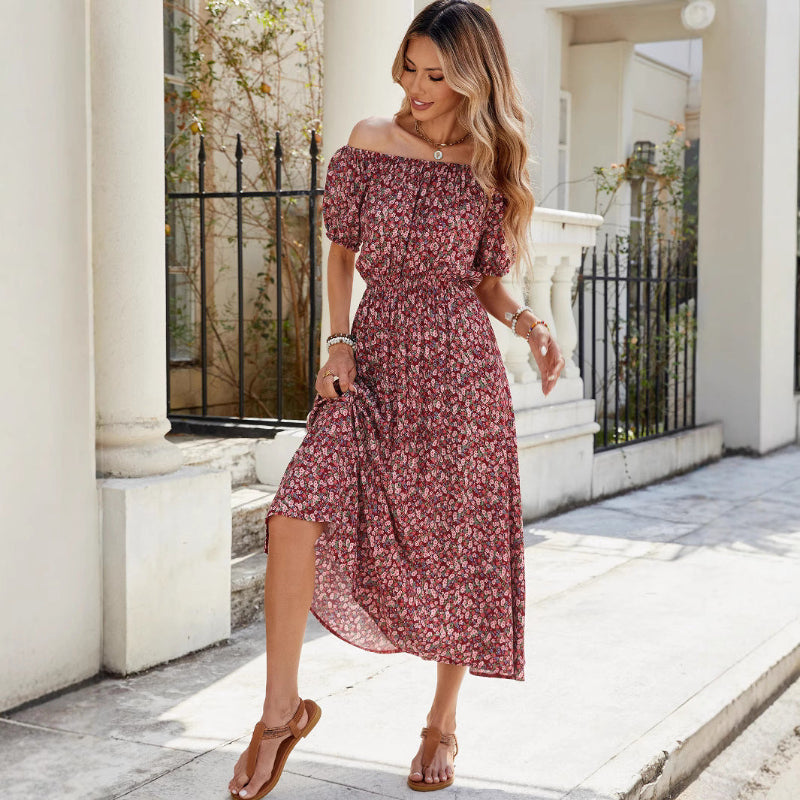 A full-length view of a person wearing a wine red, floral-printed, one-shoulder midi dress. The dress has a pleated A-line silhouette, an elastic waist, and a single short sleeve.