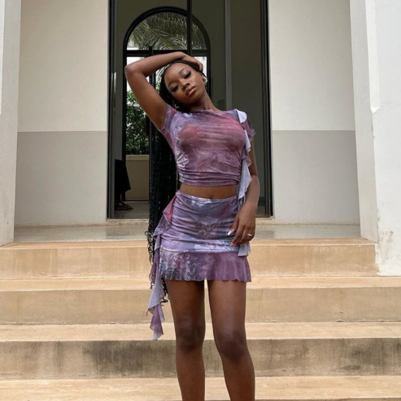 A woman wearing a purple fitted dress with a ruffle hem short top and matching mini skirt set, posing confidently.
