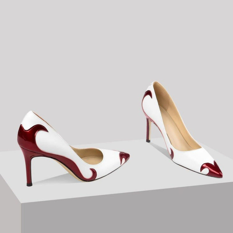 A close-up of a black satin high heel shoe with a pointed toe and a slender heel, featuring an embroidered white skull pattern and a dragonfly embellishment on the side.