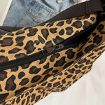 Close-up of a leopard print canvas tote bag with an adjustable strap, zipper closure, and visible interior pockets, displayed in a casual setting.