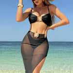 A model wearing a three-piece swimsuit set, which includes a bikini top, bikini bottoms, and a long mesh cover-up dress. The swimsuit is available in various colors and is shown in a beach setting.