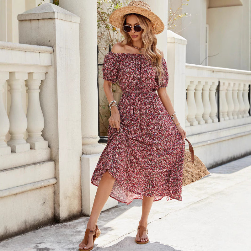 A full-length view of a person wearing a wine red, floral-printed, one-shoulder midi dress. The dress has a pleated A-line silhouette, an elastic waist, and a single short sleeve.