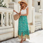 A high-waisted midi skirt with a bold digital print and a daring side slit, shown in a dark blue color. The skirt has snap buttons and is styled with a simple top.