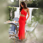 A woman wearing a sleeveless, solid red midi dress with an oblique collar. The dress has a long, flowing skirt, and she is standing in a resort-style setting.