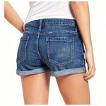 A model wearing deep blue, high-waisted denim shorts with a cuffed hem, paired with a white t-shirt. The frayed edges of the shorts are visible, and the overall style is casual and chic.