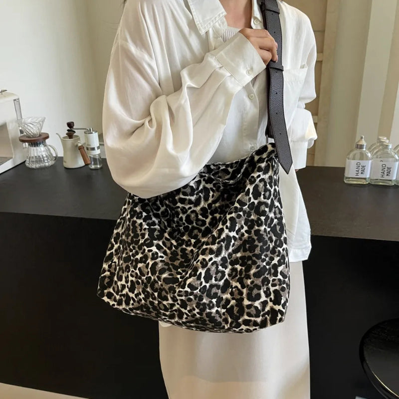 Close-up of a leopard print canvas tote bag with an adjustable strap, zipper closure, and visible interior pockets, displayed in a casual setting.