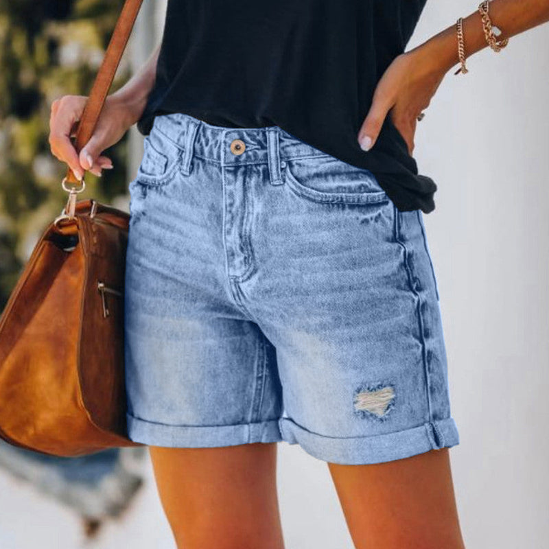 A pair of blue, high-waisted fringed denim shorts with distressed details. The shorts are laid flat on a neutral background, showcasing the style and texture. The fringe detailing is prominent along the hemline.