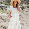 Woman in a white dress and straw hat standing on a sandy beach.